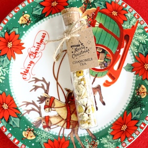 May include: A clear glass vial filled with chamomile tea, sealed with a cork and tied with twine. A small tag reads "Have a Merry Christmas" and "Chamomile Tea." The vial rests on a Christmas-themed plate with reindeer and poinsettias.