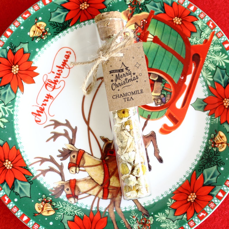 May include: A clear glass vial filled with chamomile tea, sealed with a cork and tied with twine. A small tag reads "Have a Merry Christmas" and "Chamomile Tea." The vial rests on a Christmas-themed plate with reindeer and poinsettias.