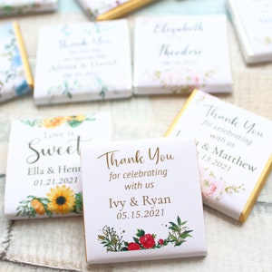 May include: A set of small, white chocolate bars with floral designs and personalized wedding messages. The bars feature gold foil accents and text that reads "Love is Sweet", "Thank You for celebrating with us Ivy & Ryan 05.15.2021", and "Thank You for celebrating with us Ella & Henry 01.21.2021".