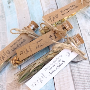 Könnte beinhalten: Vier kleine Glasflaschen mit Korkverschlüssen, gefüllt mit getrockneten Kräutern und Blumen. Jede Flasche hat ein braunes Papieretikett mit handschriftlichem Text, darunter die Namen "Andrea & Gabrielle" und das Datum "18. August 2021".