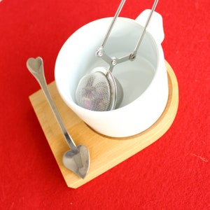 May include: A white ceramic teacup with a stainless steel tea infuser and a heart-shaped spoon, resting on a wooden coaster. The tea infuser is a mesh ball held by a handle. The spoon is silver. The background is red.