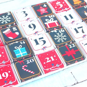 May include: Advent calendar with 24 numbered squares. Each square has a red or black background with Christmas-themed illustrations like snowflakes, gifts, and a stocking. Numbers 1-23 are visible, with a bell on the 24th square.