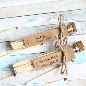 Könnte beinhalten: Zwei Glasröhrchen gefüllt mit Streichhölzern, gebunden mit Schnur und einem braunen Papieretikett. Ein Etikett lautet "Danke! marissa david" und das andere "The Perfect Match Andrea & Gabrielle".