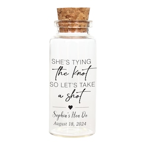 Puede incluir: Una botella de vidrio transparente con tapón de corcho. La botella tiene el texto "SHE'S TYING the knot SO LET'S TAKE a shot Sophia's Hen Do August 18, 2024" en escritura negra. La botella es probablemente para una boda o despedida de soltera.