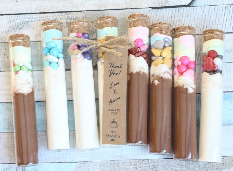 May include: Eight glass test tubes filled with different colored candies and powders, tied with twine and a brown paper tag that reads "Thank You! Zena & Amino March 12, 2023 Hot Chocolate Mix".