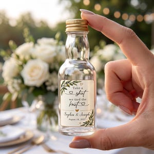 May include: A small clear glass bottle with a gold screw cap featuring a rectangular label with olive branch illustrations and the text: Take a shot we tied the knot, Sophia & Joseph, August 18, 2027, decorated with small heart icons.