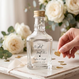 Könnte beinhalten: Eine klare Glasflasche mit goldenem Schraubverschluss und dem schwarzen Aufdruck Take a shot we tied the knot, Sophia & Joseph sowie August 18, 2026. Die Flasche hat einen quadratischen Boden und einen gerippten Flaschenhals als Dekoration.