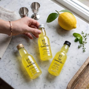 May include: Three small glass bottles filled with yellow limoncello, each featuring a label that reads Limoncello, Melinda's Hen Party, March 11, 2026. The bottles have metallic caps and are arranged near a whole yellow lemon and two metal shot glasses.