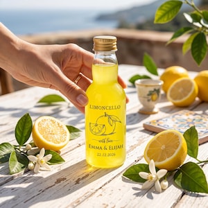 May include: A clear glass bottle filled with bright yellow limoncello, featuring a gold cap and a label printed with a lemon illustration, the text "LIMONCELLO", "with love", "EMMA & ELIJAH", and the date "22.12.2026" in black ink.