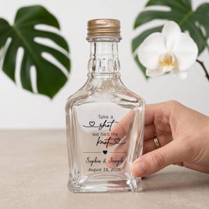 May include: A clear glass bottle with a gold-coloured screw cap, featuring black printed text that reads: Take a shot we tied the knot, followed by the names Sophia & Joseph and the date August 18, 2026, with small heart graphic accents.