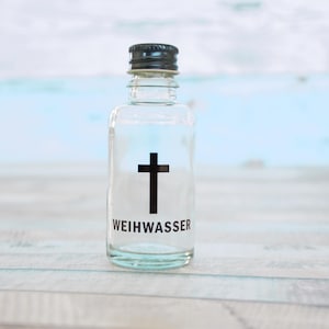 Pode incluir: Uma garrafa de vidro transparente com uma cruz preta e a palavra "WEIHWASSER" impressa. A garrafa tem uma tampa preta e é preenchida com um líquido transparente. O fundo é azul claro e branco.