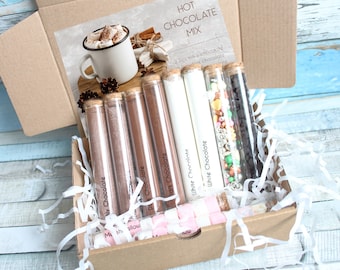 May include: A brown cardboard box filled with white tissue paper and glass test tubes containing hot chocolate mix, marshmallows, and sprinkles. The box has a label that reads "HOT CHOCOLATE MIX" and "2 Pcs milky chocolate".