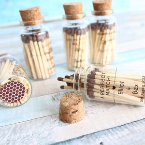 以下が含まれることがあります： 木製のマッチが詰まった小さなガラス瓶が4つあります。瓶にはコルクの蓋が付いており、パーソナライズされたテキストでラベル付けされています。1つの瓶のテキストには、ハートのシンボルと「Elliot & Bex」という名前、そして「26th August 2023」という日付が書かれています。