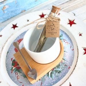 May include: A Christmas-themed gift set featuring olive leaf tea. The set includes a glass vial with a cork stopper, a white ceramic teacup, a wooden coaster, and a heart-shaped spoon. A tag reads "Christmas Tea, Olive Leaf Tea."