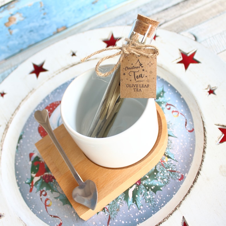 May include: A Christmas-themed gift set featuring olive leaf tea. The set includes a glass vial with a cork stopper, a white ceramic teacup, a wooden coaster, and a heart-shaped spoon. A tag reads "Christmas Tea, Olive Leaf Tea."