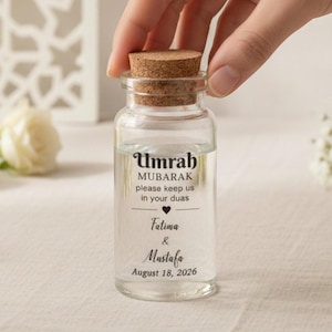 May include: A clear glass bottle with a cork stopper. The bottle contains a clear liquid and has text that reads "Umrah MUBARAK please keep us in your duas Fatima & Mustafa August 18, 2026."