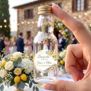 May include: A small, clear glass bottle with a gold screw cap, featuring a circular label with a watercolor illustration of lemons and a coastal town. The text on the label reads Limoncello with love Grace & Ethan Le 16 août 2026.