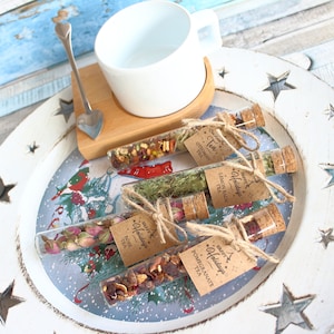 May include: A white teacup and heart-shaped spoon rest on a wooden coaster. Four glass tubes filled with loose leaf tea are displayed on a decorative tray. Each tube has a cork stopper and a tag that reads "Happy Holidays Tea".