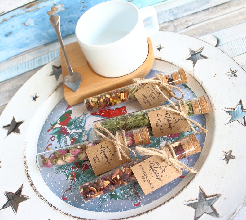 May include: A white teacup and heart-shaped spoon rest on a wooden coaster. Four glass tubes filled with loose leaf tea are displayed on a decorative tray. Each tube has a cork stopper and a tag that reads "Happy Holidays Tea".