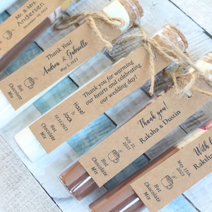 May include: Assortment of small glass bottles filled with hot chocolate mix, tied with twine and personalized with a brown paper tag. Each tag includes the couple's names and wedding date. The text on the tags reads: "Thank You! Andrea & Gabrielle May 5, 2023", "Thank you for warming our hearts and celebrating our wedding day! Hazel & Jack 10.10.2023", "Thank you! Raksha & Dustin July 21, 2023", and "With love Raksha & Dustin May 15th, 2023".