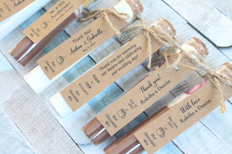 May include: Assortment of small glass bottles filled with hot chocolate mix, tied with twine and personalized with a brown paper tag. Each tag includes the couple's names and wedding date. The text on the tags reads: "Thank You! Andrea & Gabrielle May 5, 2023", "Thank you for warming our hearts and celebrating our wedding day! Hazel & Jack 10.10.2023", "Thank you! Raksha & Dustin July 21, 2023", and "With love Raksha & Dustin May 15th, 2023".