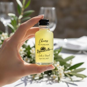 May include: A small clear glass bottle filled with yellow liquid, featuring a black screw cap. The label displays the text Cheers TO FOREVER Limoncello Leila & Darrel April 24, 2027, with a graphic of a lemon slice above the word Limoncello.