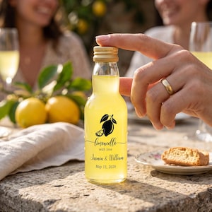 May include: A small clear glass bottle filled with yellow liquid, featuring a gold cap and a black graphic of a lemon. The label text reads: Limoncello with love, Jasmin & William, May 15, 2026. The bottle has a narrow neck and a rounded base.