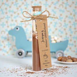 May include: A clear glass tube filled with hot chocolate mix, marshmallows, and other ingredients, tied with twine and a tag that reads "Jeniya's Winter ONEderland." The tag also includes the date 12.12.2025 and a hot chocolate graphic.