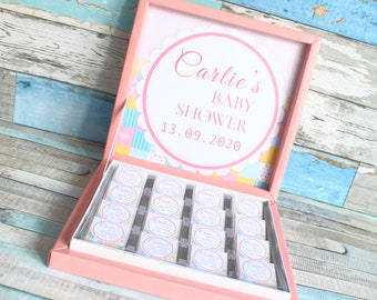 May include: A pink gift box containing multiple small square chocolates, each wrapped in paper featuring the text Carlie's BABY SHOWER 13.09.2020. The inside lid displays a large circular label with the same text and a colorful checkered pattern.