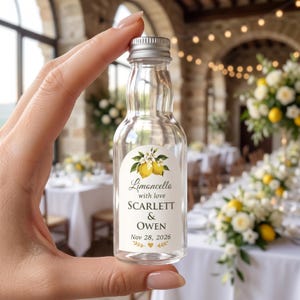 May include: A small, clear glass bottle with a silver screw cap featuring a label with a lemon illustration, the text Limoncello with love SCARLETT & OWEN Nov 28, 2026, and a small heart icon. The bottle has a narrow neck and a rounded body.