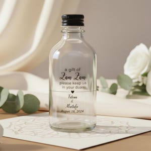 May include: A clear glass bottle with a black cap, featuring the text "a gift of Zam Zam" and a personalized message. The bottle is designed for a special occasion, with the names Fatima & Mustafo and the date August 18, 2024.