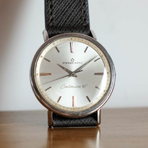 May include: A silver wristwatch with a white face and black leather band. The watch face has black hands and black Roman numeral hour markers. The watch face also has the text "Eterna-Matic" and "Centenarie '61".