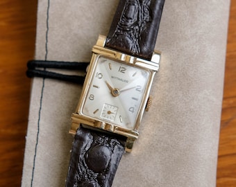 May include: A gold-tone wristwatch with a white face and black leather strap. The watch face has black Roman numeral hour markers and the text "WITTNAUER" printed on it.