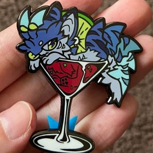 以下が含まれることがあります： 青いストライプのシャツを着て、青と白のストライプの尻尾を持つ、カートゥーンの猫が赤い液体とライムのくさびが入ったマティーニグラスを持っている、青と白のエナメルピン。