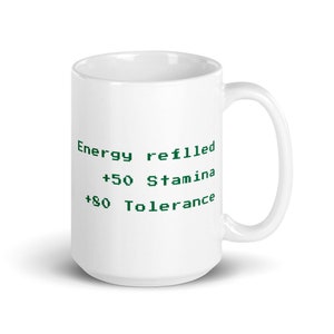 May include: White ceramic mug with a curved handle. The mug features green pixelated text that reads "Energy refilled +50 Stamina +80 Tolerance". The mug is designed for hot or cold beverages.