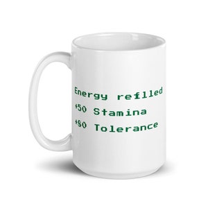 May include: White ceramic mug with a curved handle. The mug features green text that reads "Energy refilled, +50 Stamina, +80 Tolerance". The mug is designed for hot or cold beverages.