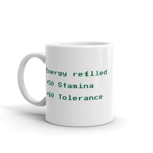 May include: White ceramic mug with a curved handle. The mug features green text that reads "Energy refilled", "+50 Stamina", and "+80 Tolerance". A simple, everyday coffee mug.