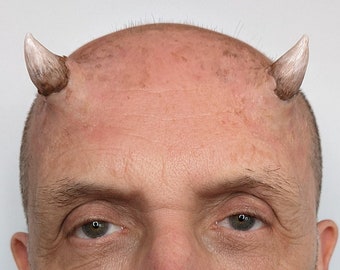May include: A close-up portrait of a person with short, brown hair and a goatee. The person is wearing a black shirt and has two small, white horns attached to their forehead.
