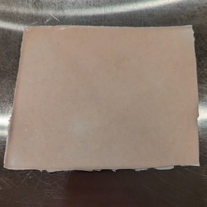 May include: A rectangular piece of light pink material with a slightly uneven edge. The material has a smooth, matte surface and appears to be a solid color. The background is a metallic surface.