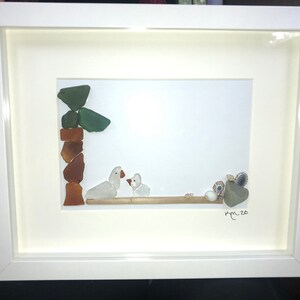 May include: Framed artwork featuring a beach scene. The art is made of sea glass and shells, depicting two white ducks on a wooden branch. The frame is white and the artwork is signed.