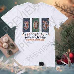 Op de afbeelding: Wit T-shirt met het kengetal "303" in een geruit ontwerp, met de tekst "Mile High City Denver, Colorado" eronder. Het ontwerp bevat lichtslingers. Het T-shirt wordt gepresenteerd met decoratieve elementen.