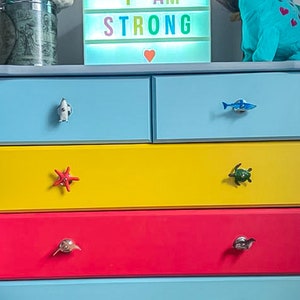 May include: A colourful chest of drawers with six drawers, each painted a different colour. The drawers have unique handles, including a starfish, a turtle, a shark, and a whale. The dresser is topped with a light-up sign that reads "I AM STRONG".