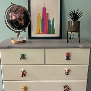 May include: A white chest of drawers with six drawers, each featuring a different character from the Super Mario Bros. video game series as a drawer pull. The chest of drawers is painted a light grey colour and has a grey top. A globe and a framed print of the New York City skyline are on top of the chest of drawers.