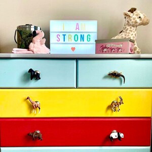 May include: A colourful chest of drawers with five drawers, each with a different animal figurine as a pull. The top drawer is light blue, the second is yellow, the third is red, the fourth is light blue, and the fifth is yellow. The dresser is decorated with a light box that says "I AM STRONG" with a pink heart.