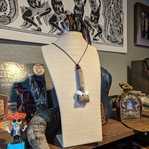 K&ouml;nnte beinhalten: Eine braune und wei&szlig;e Halskette mit einem Holzanh&auml;nger und wei&szlig;en Perlen h&auml;ngt an einer wei&szlig;en Mannequin-B&uuml;ste. Die Halskette wird auf einem Regal mit anderen Dekorationsartikeln ausgestellt.