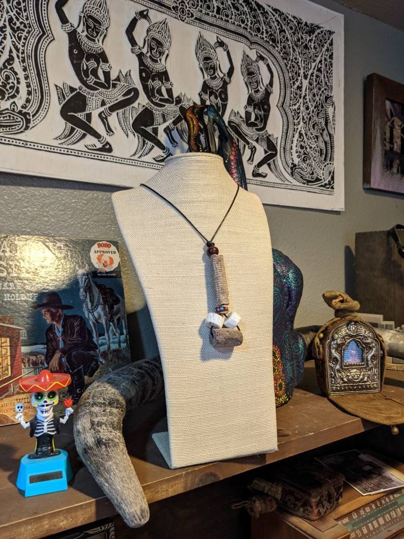 K&ouml;nnte beinhalten: Eine braune und wei&szlig;e Halskette mit einem Holzanh&auml;nger und wei&szlig;en Perlen h&auml;ngt an einer wei&szlig;en Mannequin-B&uuml;ste. Die Halskette wird auf einem Regal mit anderen Dekorationsartikeln ausgestellt.