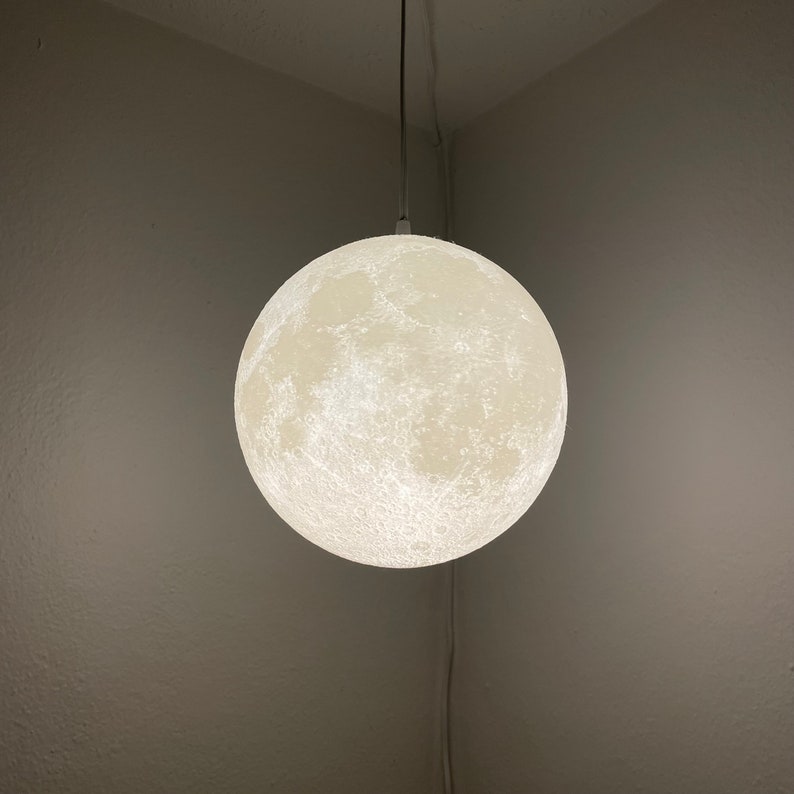 May include: A spherical, off-white pendant light resembling the moon, suspended from a thin cord. The light has a textured surface with craters and other lunar details. The light is hanging in the corner of a room with off-white walls.