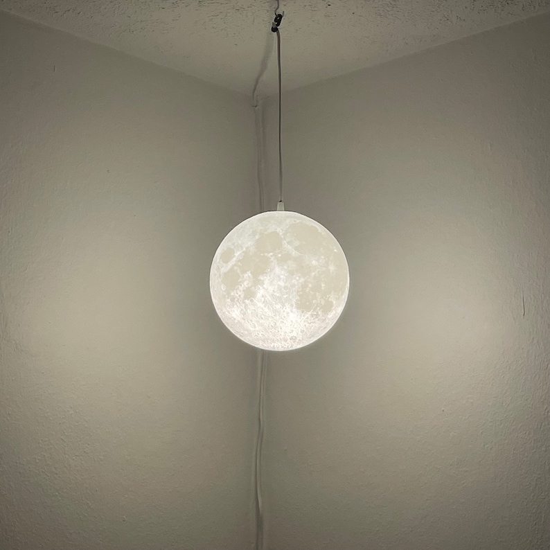 May include: A round, white moon-shaped pendant light fixture hangs from the ceiling. The light has a textured surface, resembling the moon's craters. The fixture is suspended by a thin, silver-colored cord.