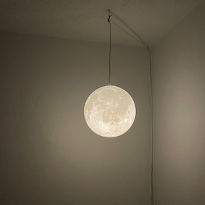 May include: A spherical, glowing moon lamp hangs from the ceiling. The lamp is white with a textured surface, mimicking the moon's craters. A thin cord and wire connect the lamp to the ceiling, casting a soft light in the room. The lamp is a decorative lighting fixture.