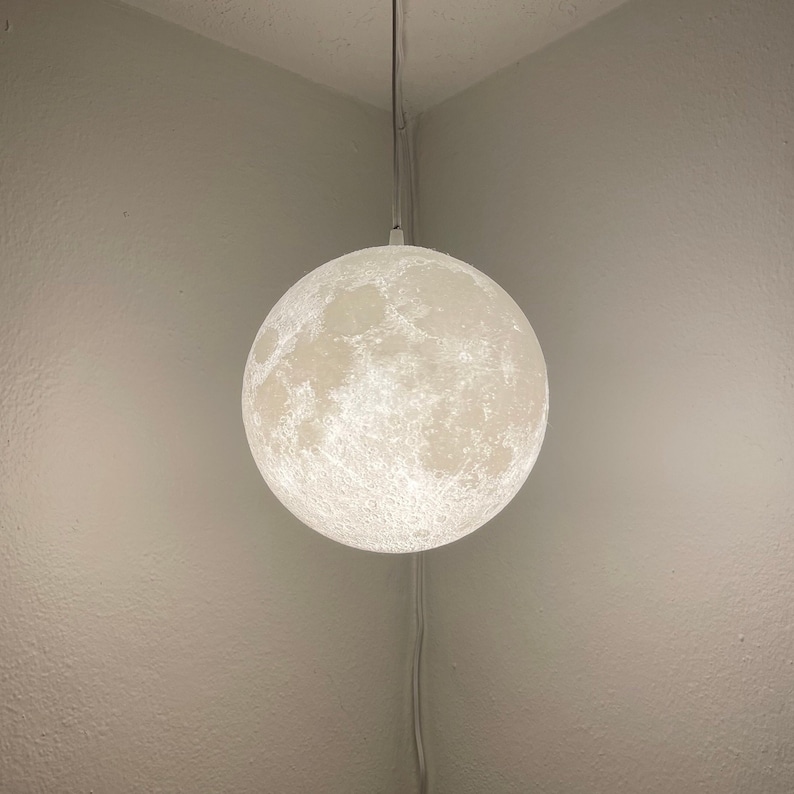 May include: A white, spherical pendant light resembling the moon, hanging from a cord. The light has a textured surface with craters and illuminated details, creating a realistic lunar appearance. The light is suspended in the corner of a room.